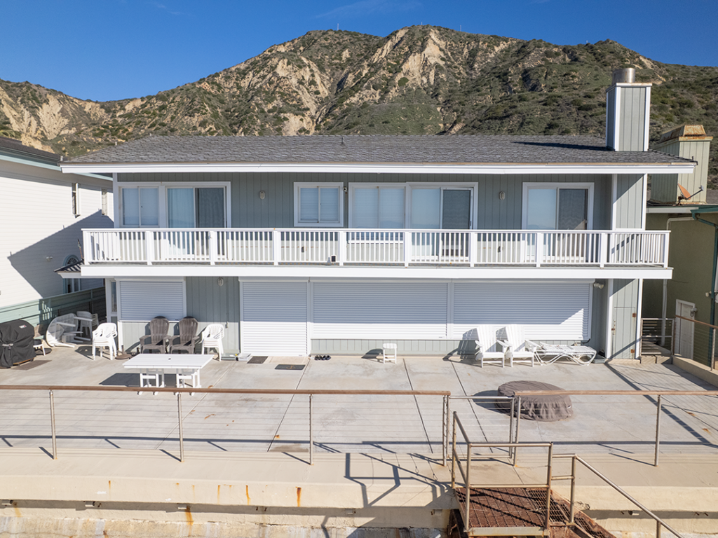 Exterior_Drone_03Web 1 A two-story beach house with a gray roof and white railing on the balcony. The structure features large windows and a patio area with lounge chairs. The coastal landscape includes mountains in the background under a clear blue sky.