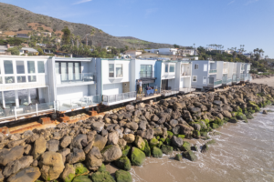 Exterior_Drone_22 A row of modern beachfront homes situated on a rocky coastline, with green moss-covered rocks in the foreground and calm ocean waves lapping at the shore. The background features a mountainous landscape under a clear blue sky.