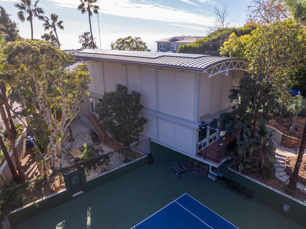 Exterior_Drone_30Web 1 Aerial view of a tennis court next to a modern building surrounded by palm trees and greenery, with ocean views in the background, showing a relaxed outdoor atmosphere.