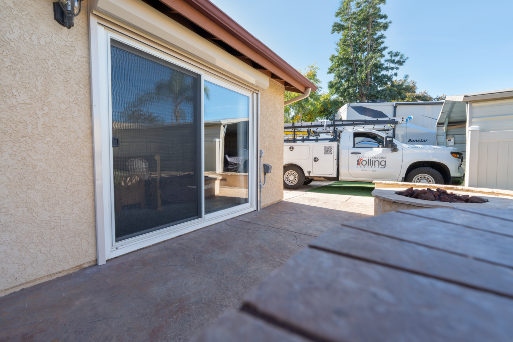 SoCal_Exterior_01 A patio view showing a sliding glass door with a screen, leading into a house. Outside, a work truck is parked nearby, with a visible logo on its side, set against a backdrop of trees and a clear blue sky.