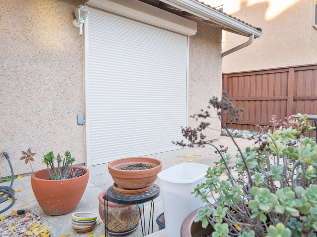 SoCal_Exterior_18Web 1 A patio area featuring a roll-up door, potted plants, and a white bucket. The wall is beige with a wooden fence in the background. The scene is outdoors with fallen leaves on the ground.