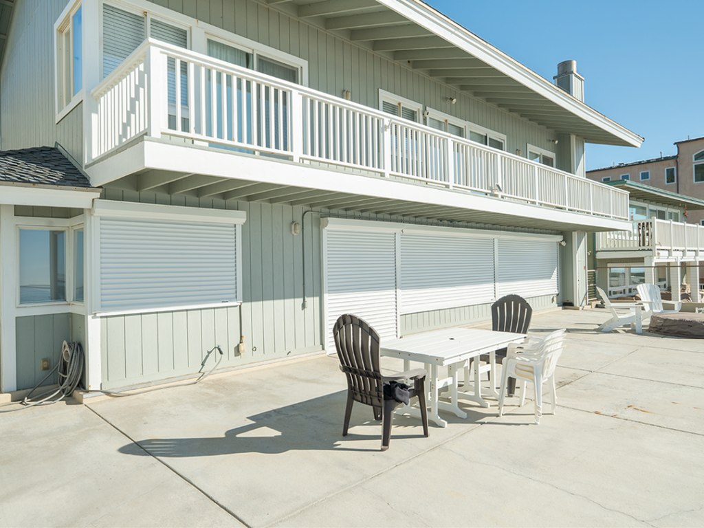 SoCal_Exterior_21Web 1 A coastal building with a balcony and white shutters, featuring a rectangular dining table surrounded by black and white chairs on a concrete patio.