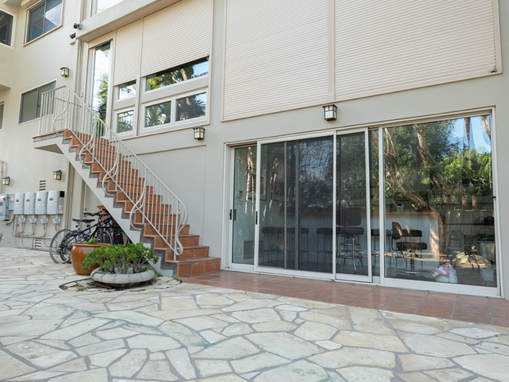 SoCal_Exterior_31Web 1 A modern building exterior with a staircase leading to a second level, large windows, and glass doors. A bike is parked near the entrance, and there are potted plants at the base of the stairs, set on a stone-paved surface.