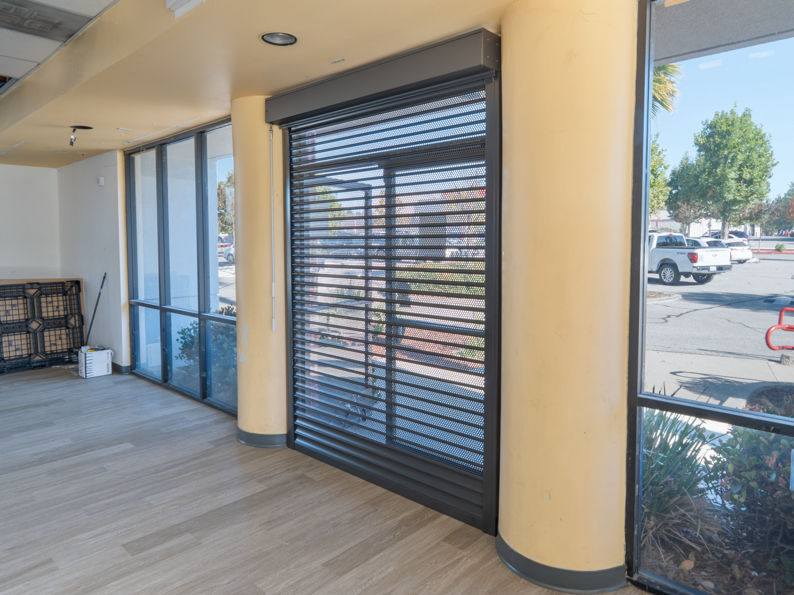 Interior view of a commercial space with a rolling security shutter and large windows allowing light. The floor is light wood, and there are two yellow columns. Outdoor greenery and a vehicle are visible through the windows.