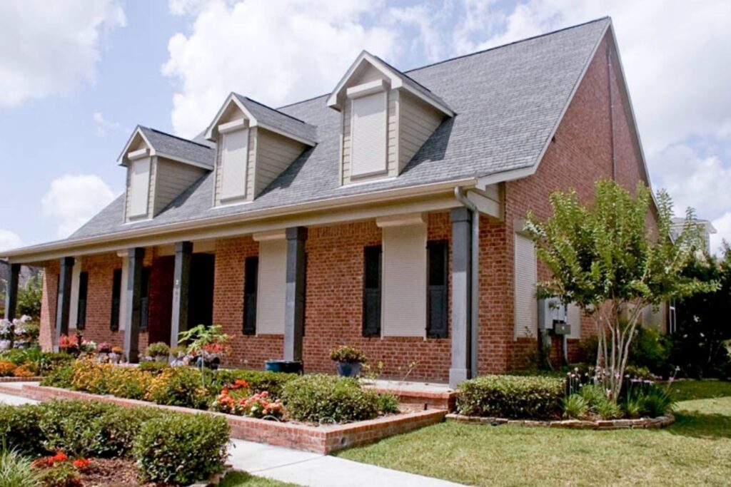 image A residential brick house with a gray roof and large front porch. The house features three gabled dormers and has flower beds with colorful plants in front. The lawn is well-maintained, and there are two trees near the entrance.
