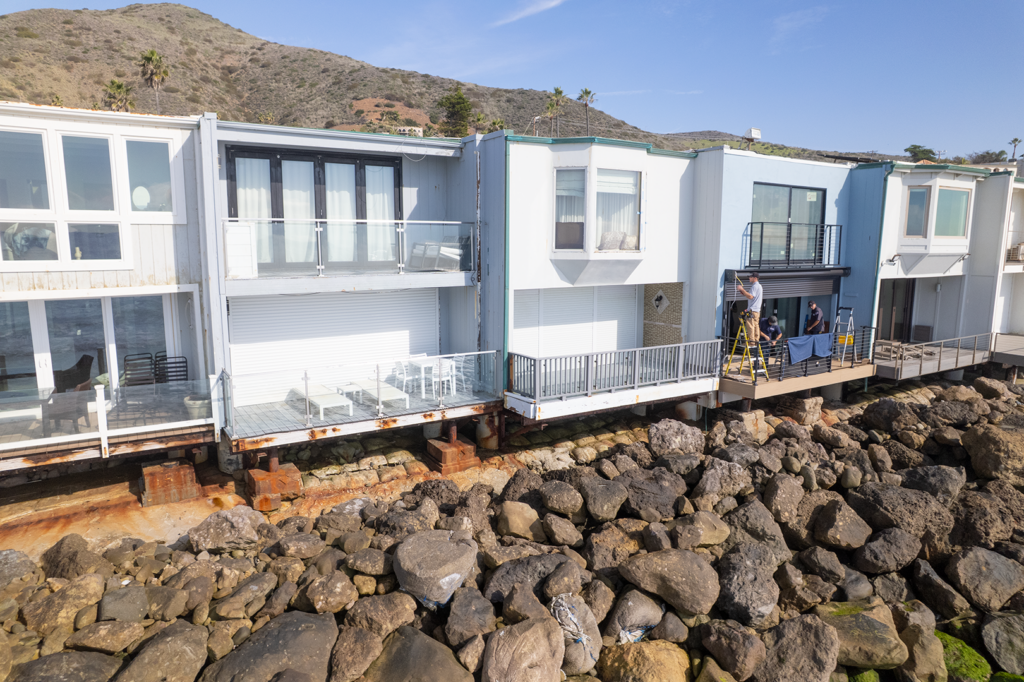 Exterior_Drone_20Web A row of beachside houses on a rocky shoreline, with one worker on a ladder making repairs. Each house features glass balconies. Behind them is a hillside and clear blue sky, with a few palm trees visible.
