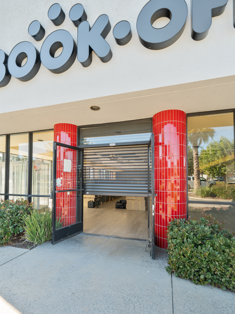 A storefront with the sign "BOOK-OP" above the entrance. The glass doors are open, revealing an interior with a tiled floor. Red columns frame the entrance, and there is landscaping with green shrubs at the base of the building.