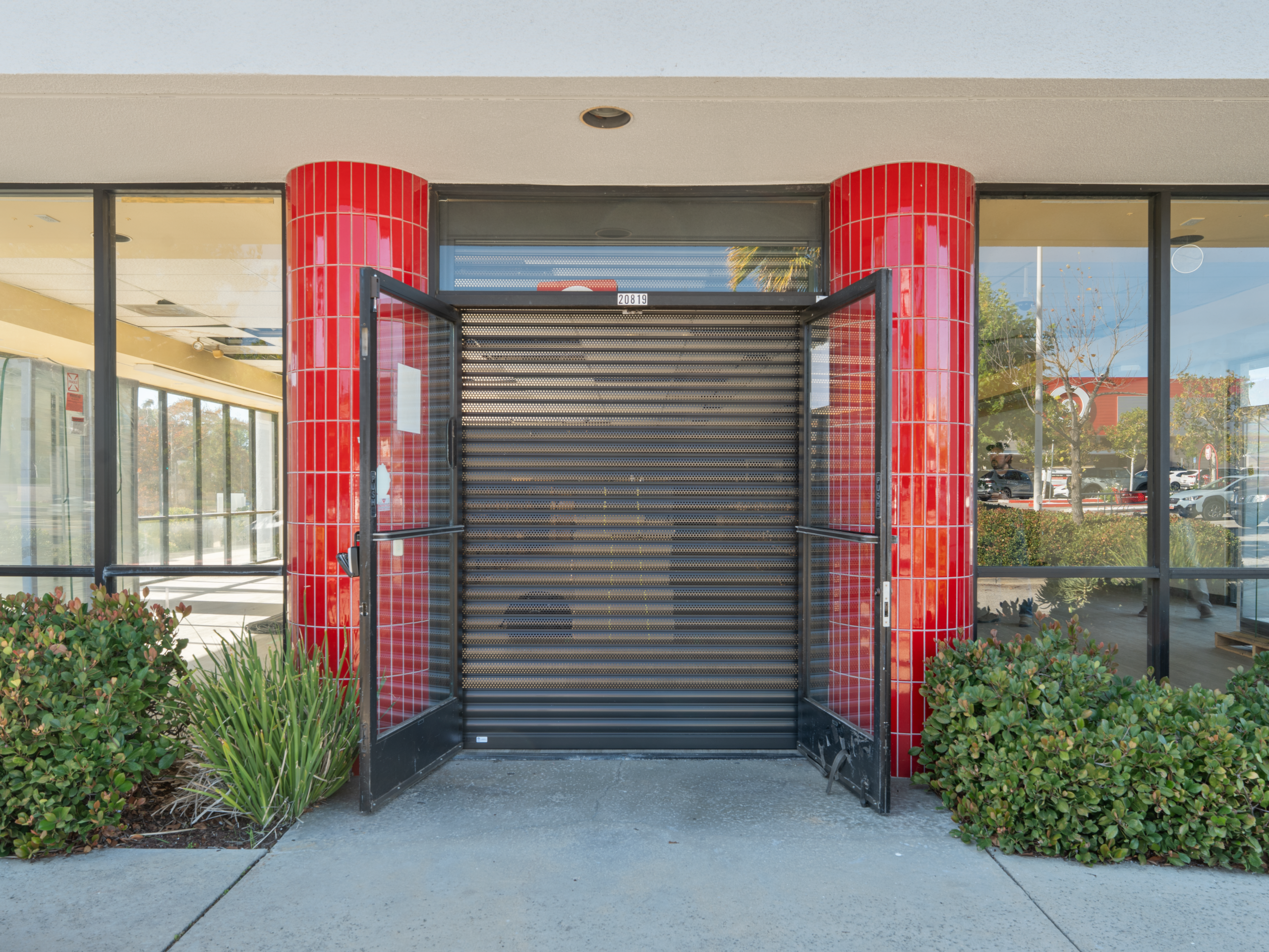A storefront with a black, rolled-down security shutter and double doors opened outward. The columns on either side are covered in red tiles. In the background, a well-maintained sidewalk and green shrubs are visible, along with a glimpse of a parking lot.