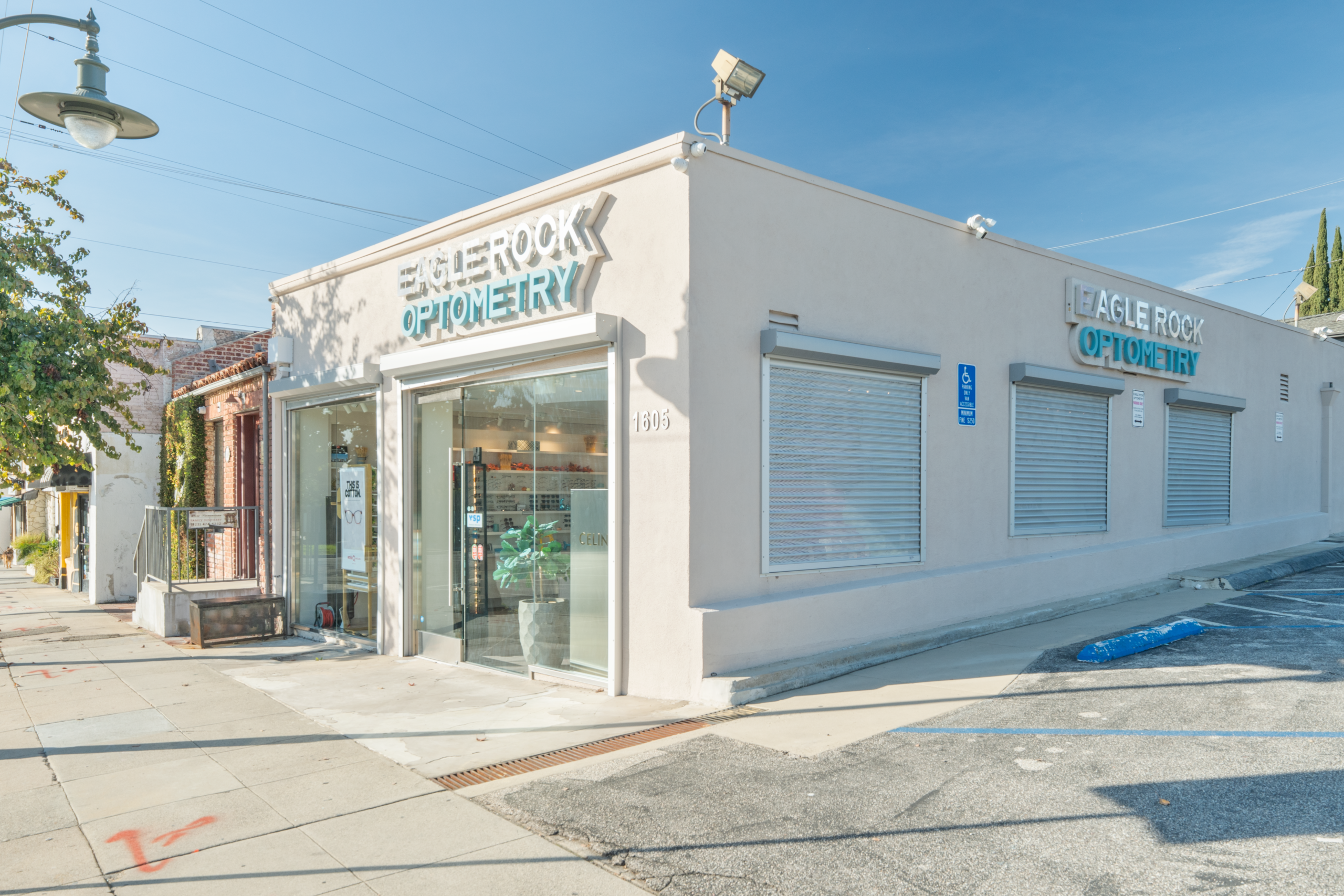 A modern optometry shop named "Eagle Rock Optometry" with large glass windows. The building features a light exterior, blue signage, and is located on a sidewalk next to a road. It has security shutters and is surrounded by greenery.