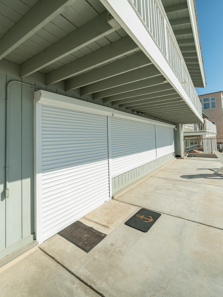 SoCal_Exterior_27Web Exterior view of a building featuring a covered patio area. The ground is concrete and a rolling white shutter is closed along a section of the wall. Two doormats are visible at the entrance, one with an anchor design.