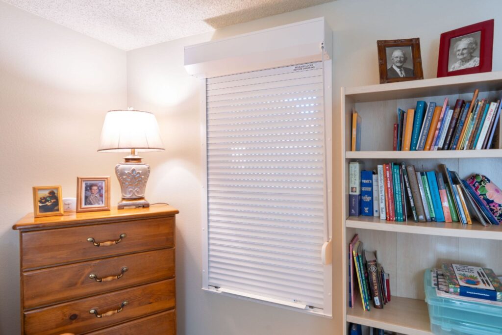 A room corner featuring a wooden dresser with framed photos on top, a lamp with a white shade, and a window with light-filtering blinds. A bookshelf nearby contains various colorful books and decorations.