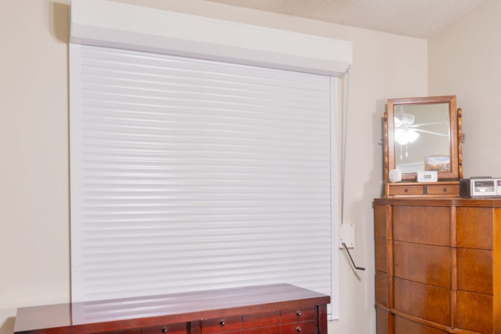 A closed white roller shade covering a window, displayed against a light-colored wall. To the right, a wooden dresser with multiple drawers and a small mirror on top reflects light in the room.