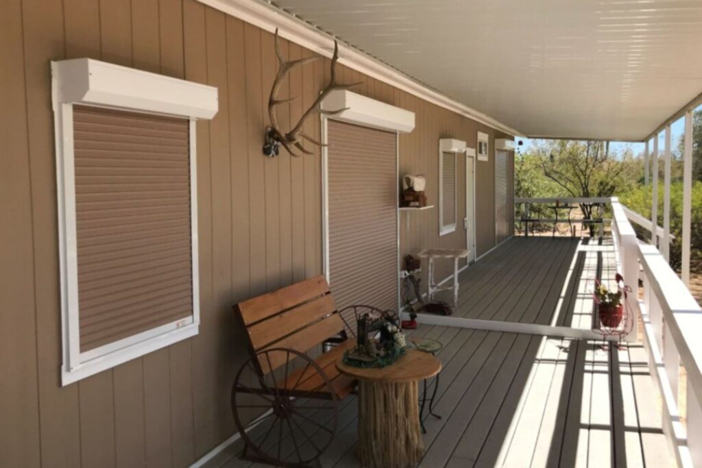 A porch of a beige house featuring closed brown window shutters, a wooden bench with a backrest, and a rustic wooden table. Decorative items, including an old bicycle wheel and a mounted pair of antlers, are positioned near the wall.