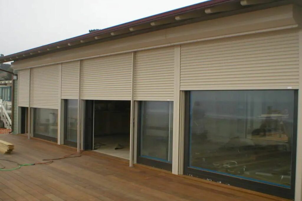 A modern building exterior with three large windows, covered by beige roller shutters. The wooden deck in front is visible, and the background shows an overcast sky.