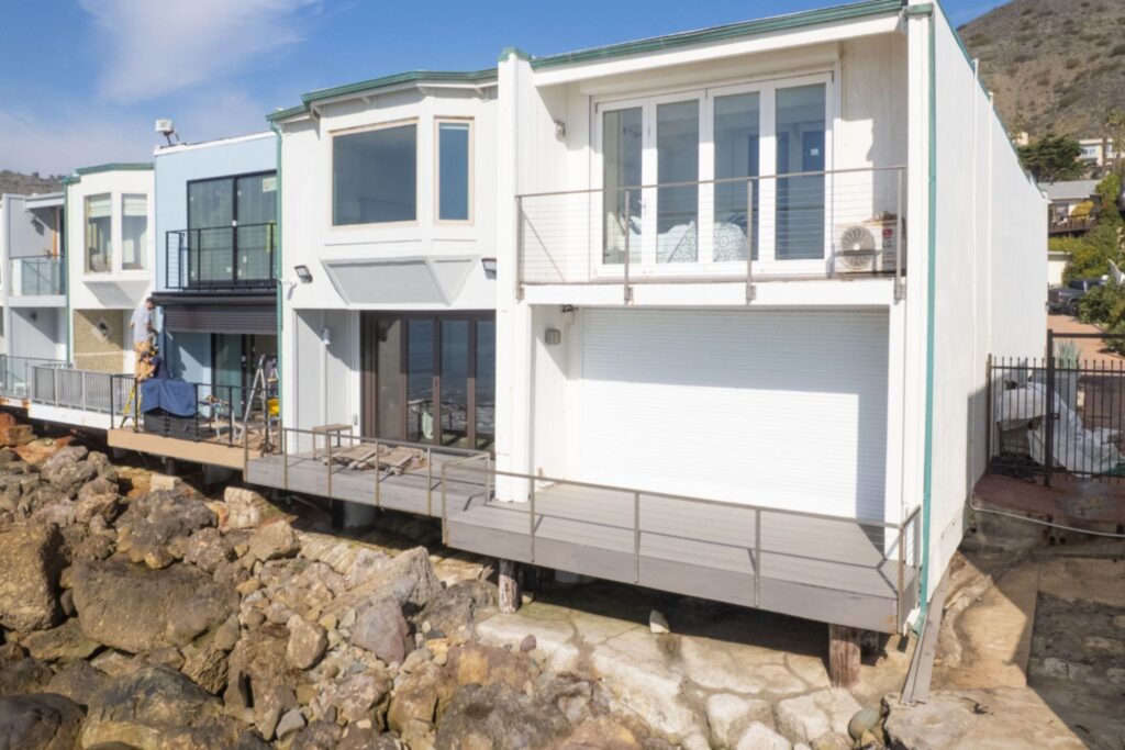 A modern waterfront house with a wooden deck extending over rocky shoreline. The structure features large windows and a white exterior, surrounded by rocky terrain and hilltop greenery in the background.