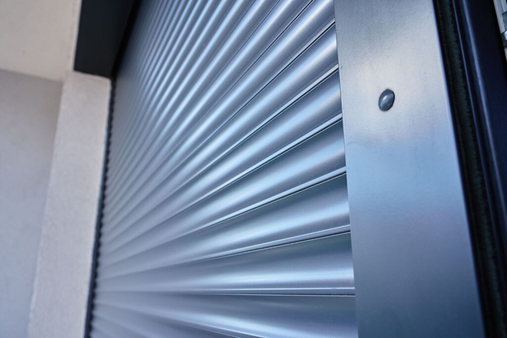 A close-up view of a gray metal rolling shutter with a textured surface, focusing on the vertical slats. The background features a light-colored wall next to the shutter.