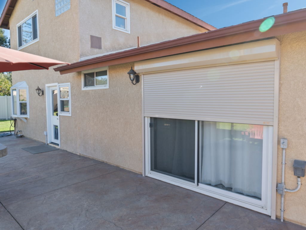 A beige stucco house with a large sliding glass door and closed white roller shutter, shown beside a concrete patio and small windows.