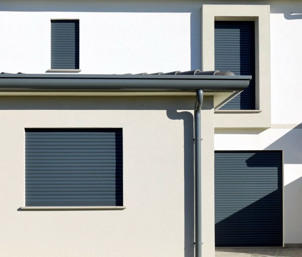 A modern white house with dark roller shutters on the windows and a gray gutter along the roofline, shown in bright sunlight.
